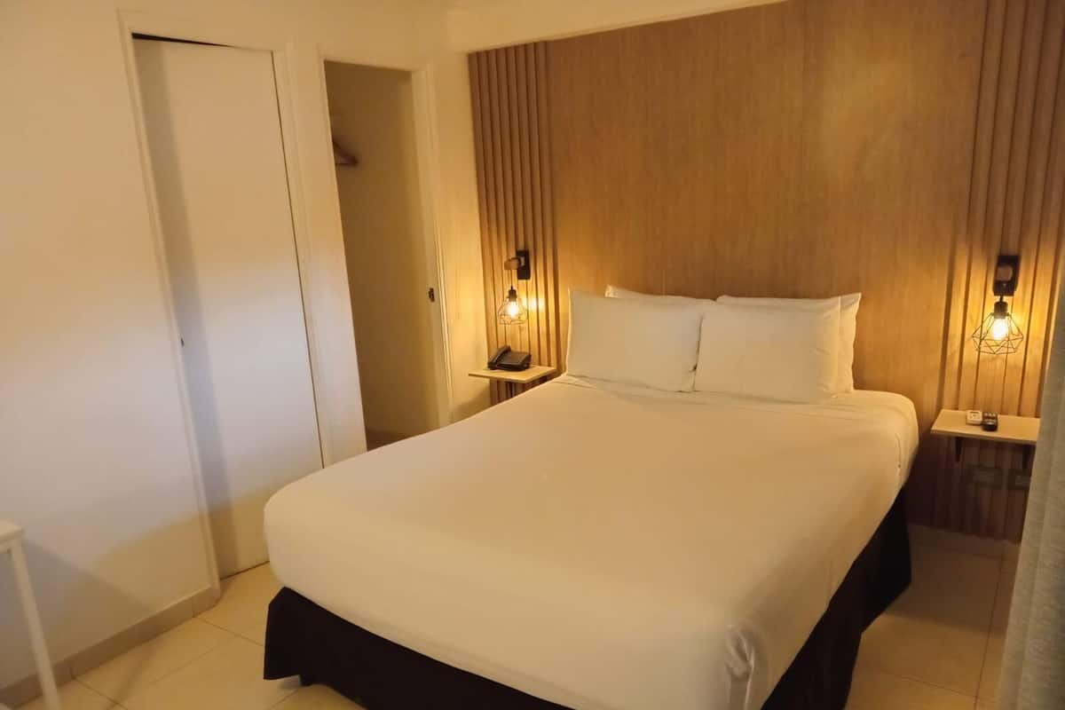 A hotel room with a large bed, bedside tables with lamps, a wooden headboard, and a door leading to another room.