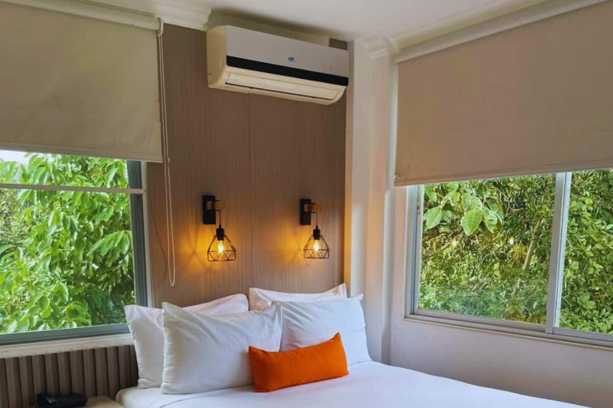 A hotel room with a large bed, two wall-mounted lights, an air conditioner, and a window with a view of greenery.