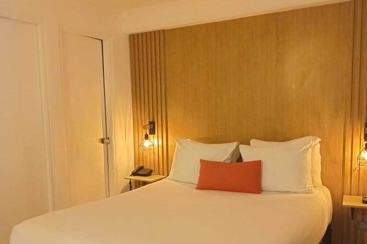 Quarto de hotel com uma cama grande, cabeceira de madeira, duas mesas de cabeceira com abajures e uma porta.