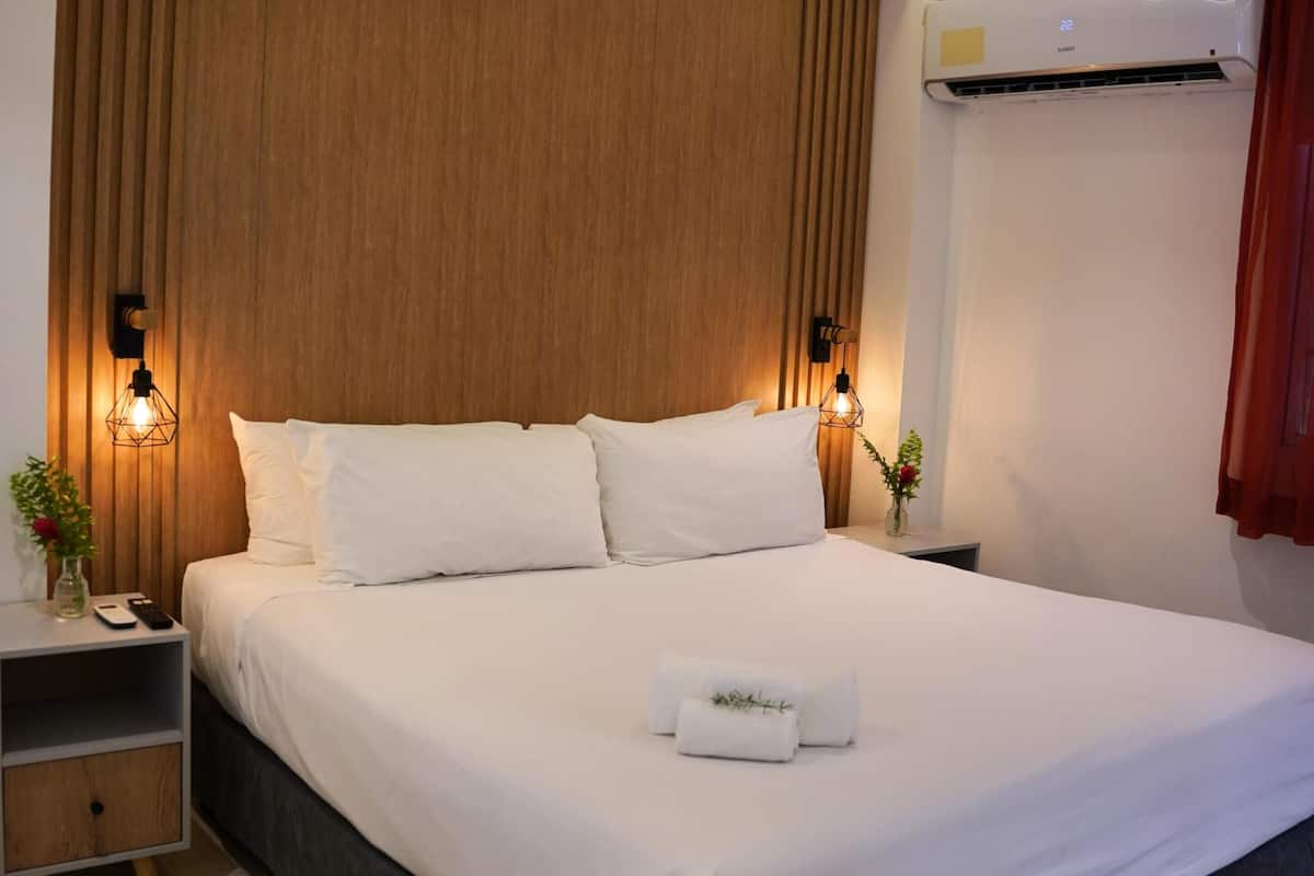 A modern hotel room with a large bed, wooden headboard, bedside lamps, a nightstand, and an air conditioning unit.