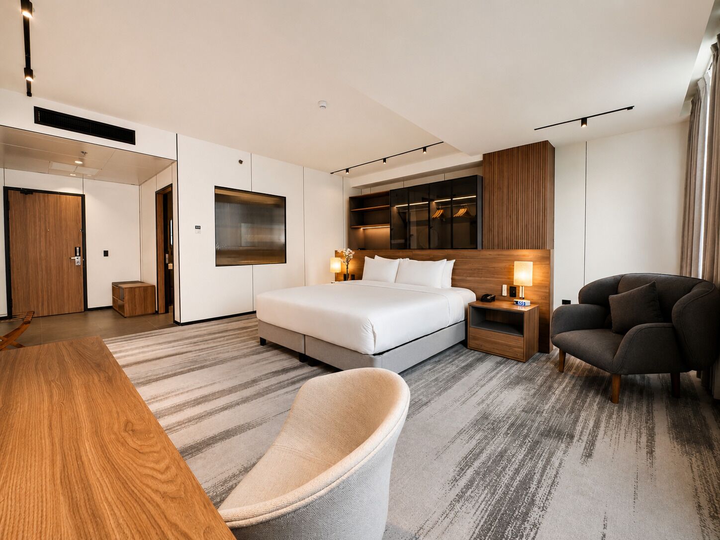 A modern hotel room with a large bed, a desk, a chair, a nightstand, and a wardrobe.