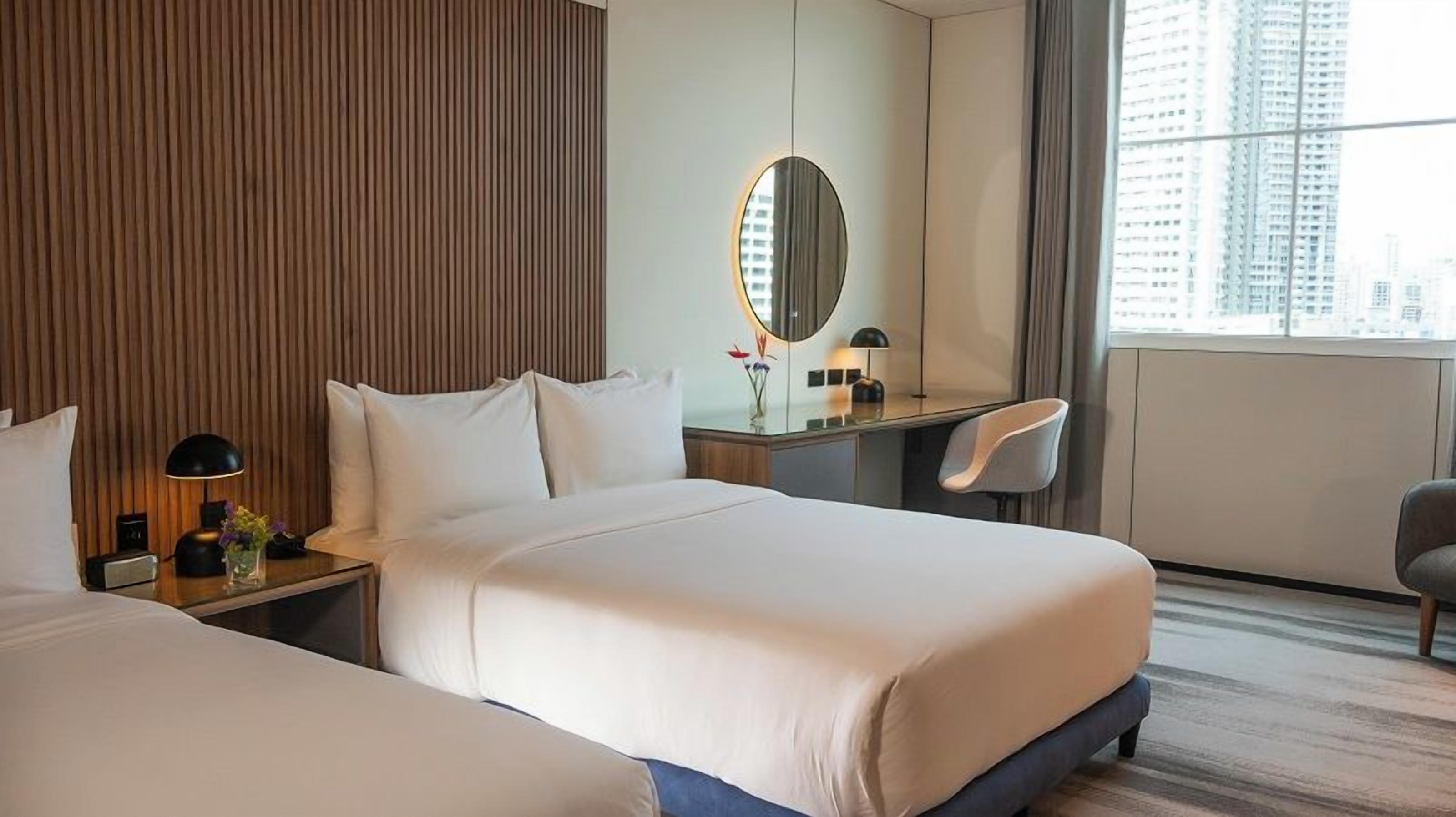 A hotel room with two beds, a desk with a chair, a large mirror, and a view of the city.