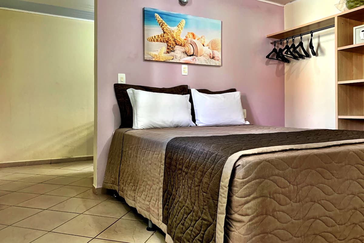 Una cama bien hecha con cubrecama acolchado, un cuadro con una estrella de mar y conchas marinas en la pared, y una estantería de madera con perchas.