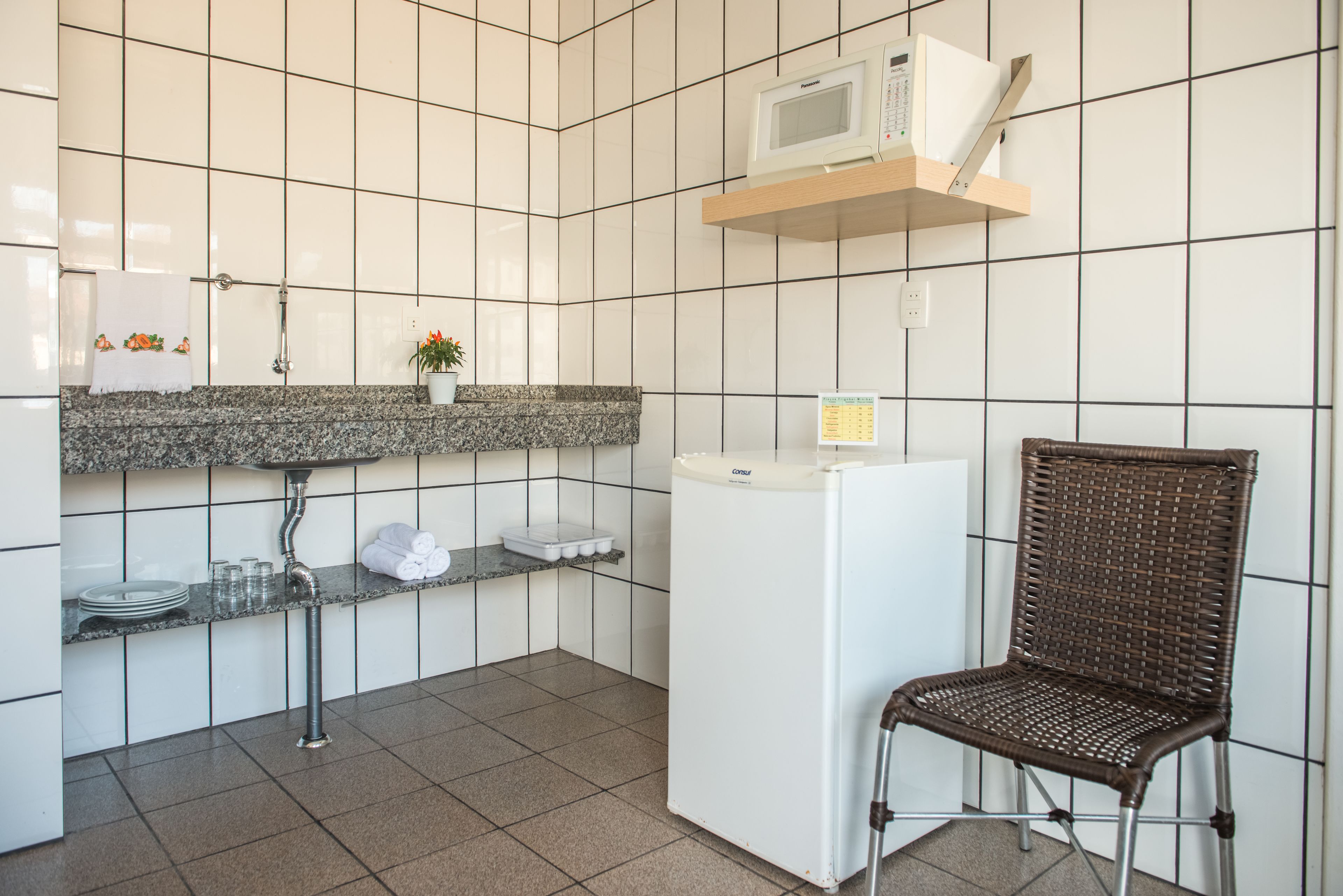 Uma cozinha com paredes revestidas de azulejos brancos, bancada de granito, geladeira branca, uma cadeira de vime e micro-ondas em uma prateleira.