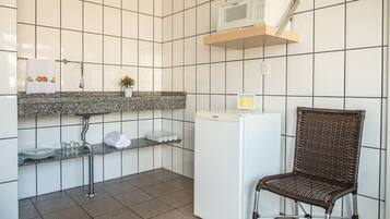 Uma cozinha com paredes revestidas de azulejos brancos, bancada de granito, geladeira branca, uma cadeira de vime e micro-ondas em uma prateleira.