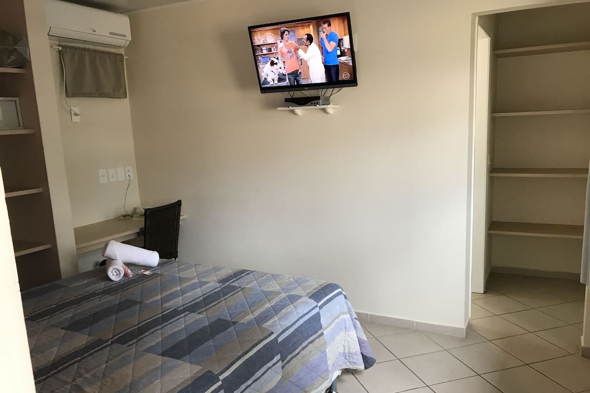 Un dormitorio con cama, televisor montado en la pared, ventana con cortina y piso de baldosas.