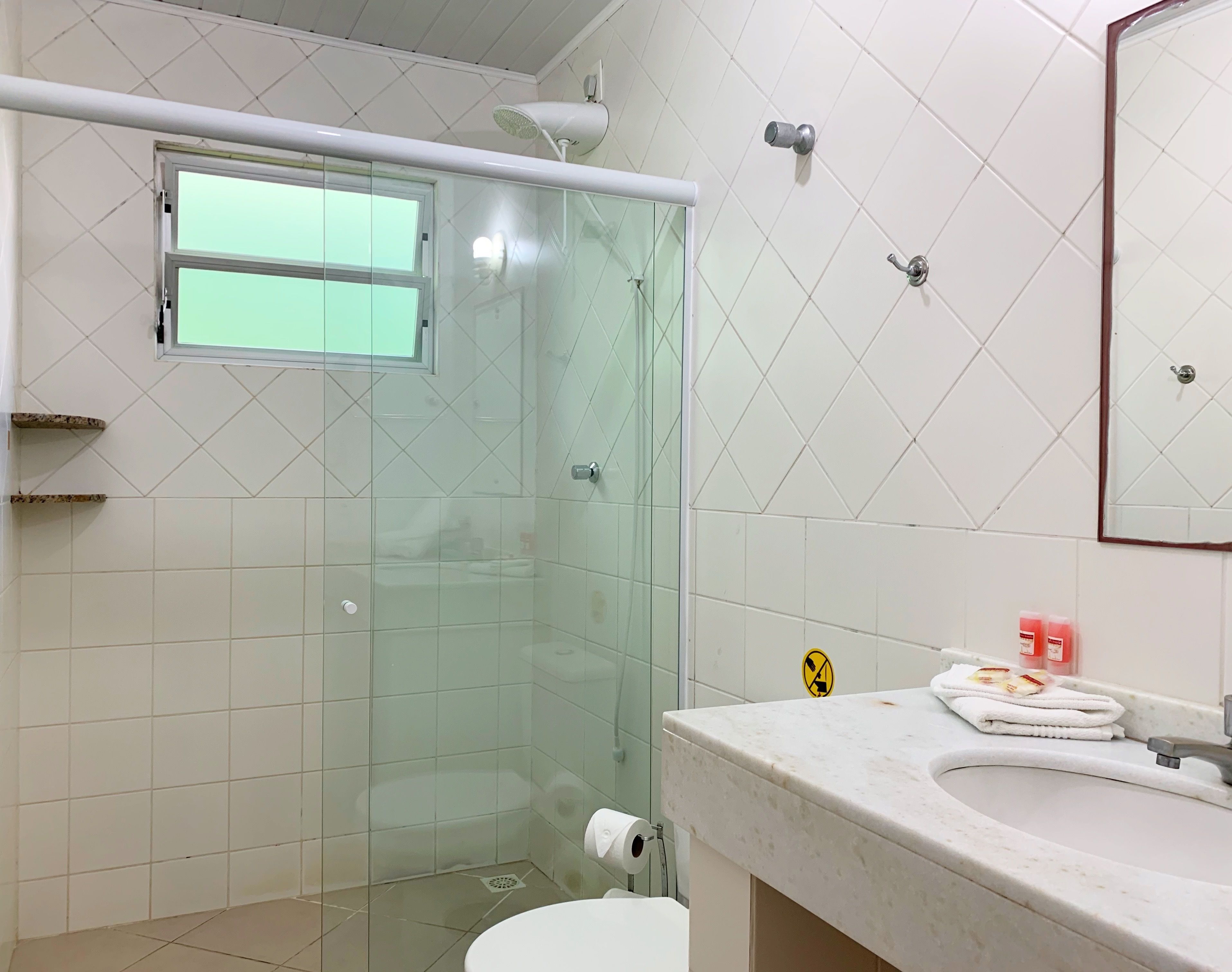 Un baño con ducha vidriada, inodoro, lavamanos con espejo y ventana sobre la ducha.