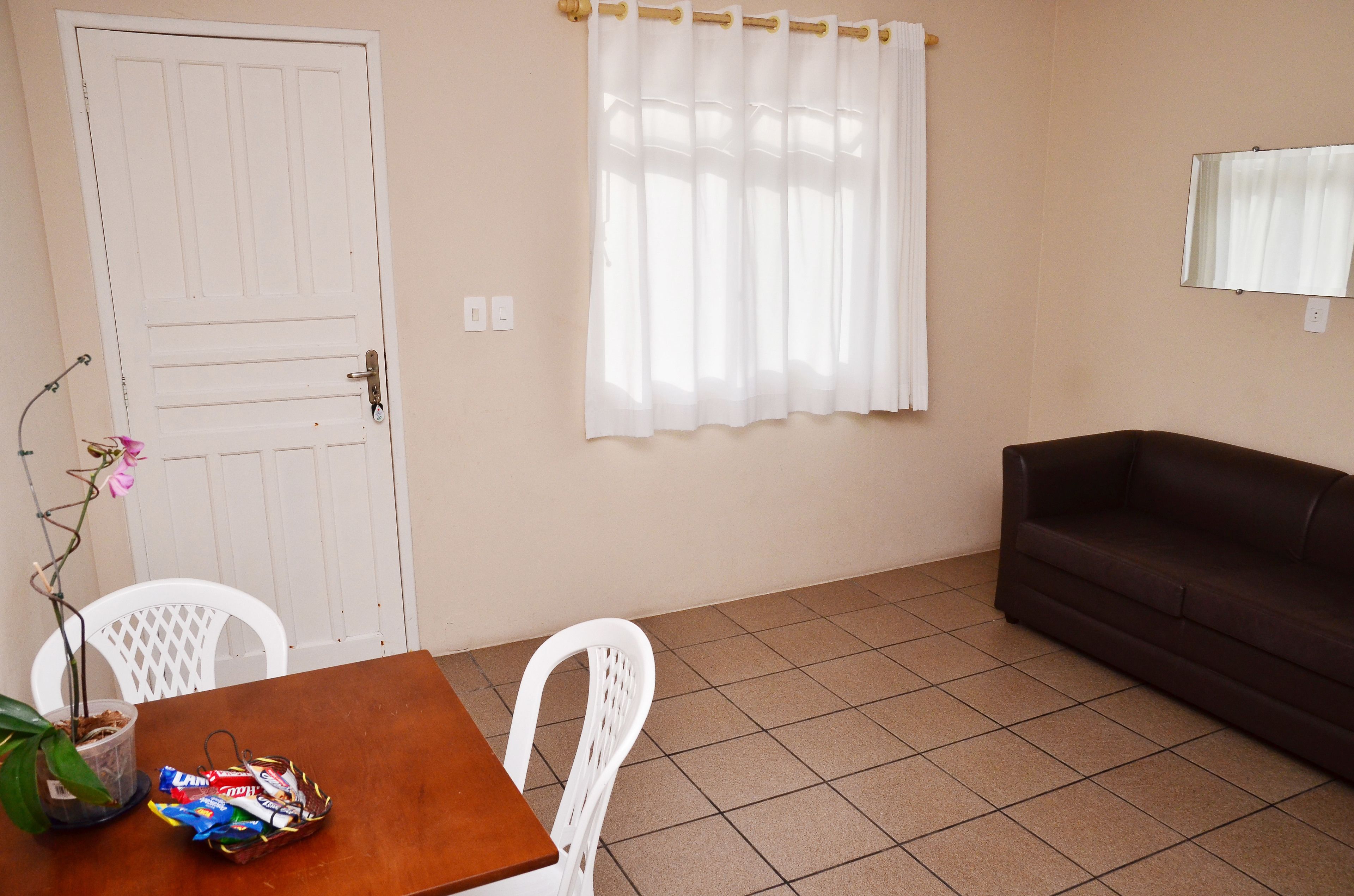 A room with a wooden table, white chairs, a brown sofa, a potted plant, and a window with white curtains.