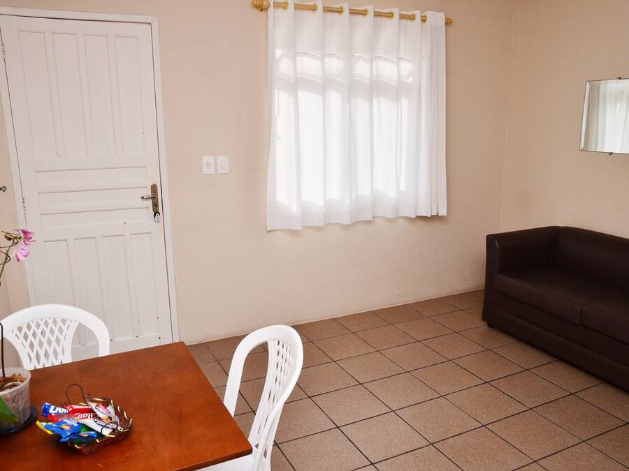 A room with a wooden table, white chairs, a brown sofa, a potted plant, and a window with white curtains.