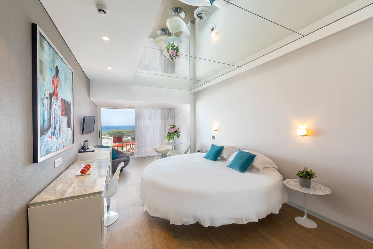 Habitación de hotel moderna con una cama grande, un escritorio con una computadora, una mesita con una planta y un balcón con vistas.