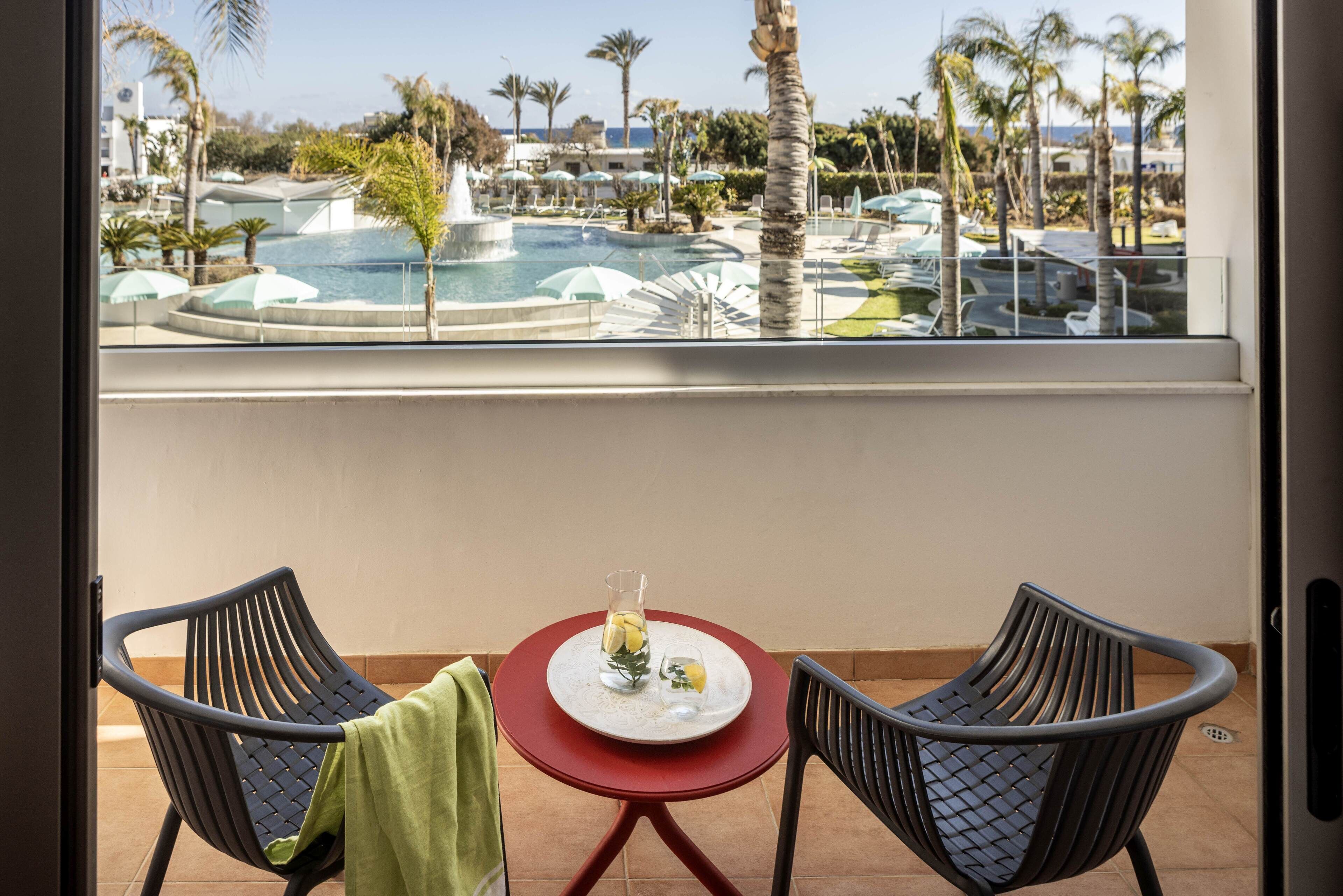 A balcony with two chairs, a small table with a glass and lemons, and a view of a pool and palm trees.