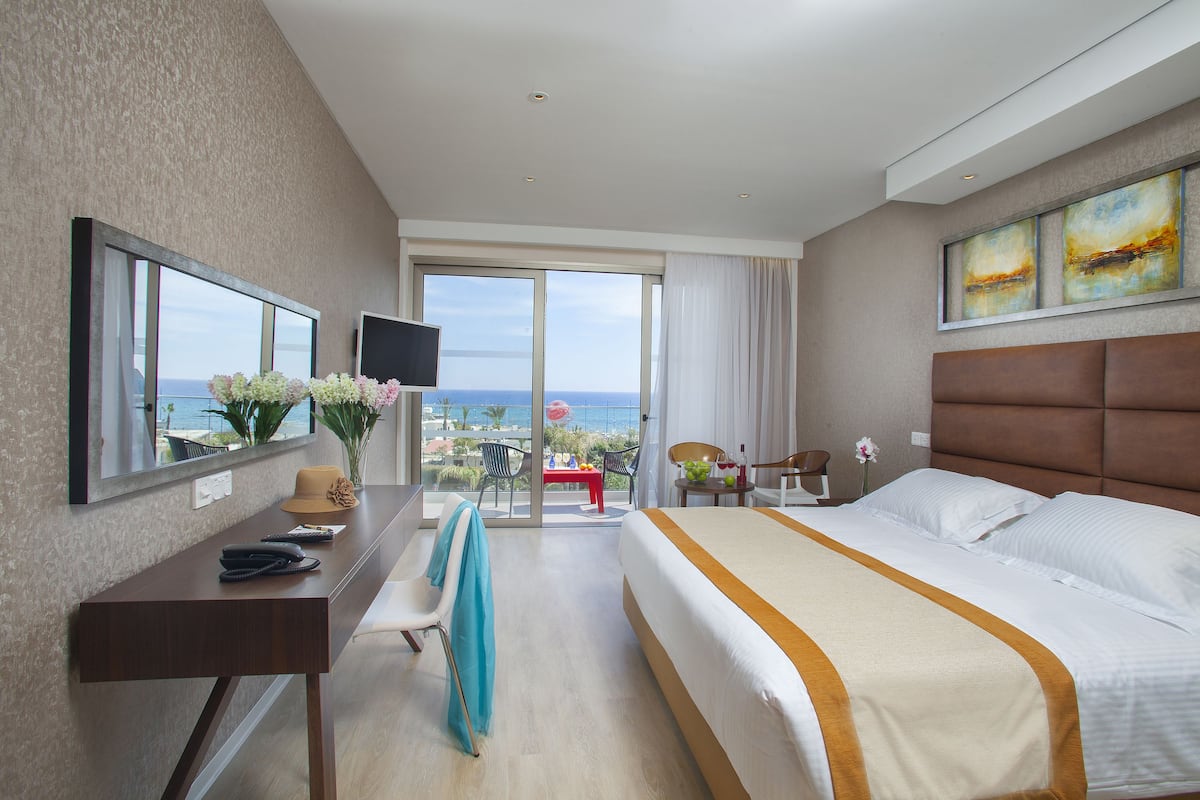 Una habitación de hotel con una cama grande, un escritorio con teléfono, una silla, un televisor y un balcón con vistas al mar.