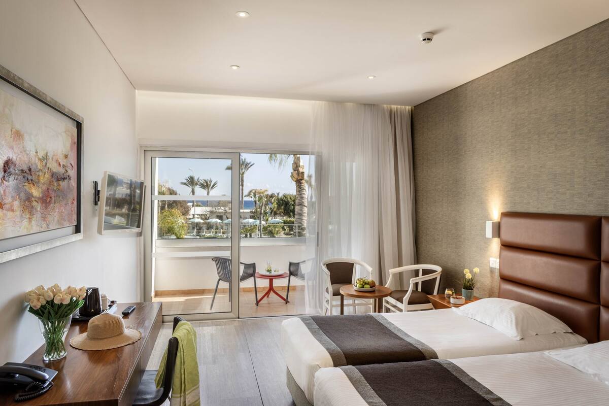 Habitación de hotel con cama, escritorio con teléfono, jarrón de flores, silla, mesita con planta y vista a palmeras a través de una puerta de cristal.