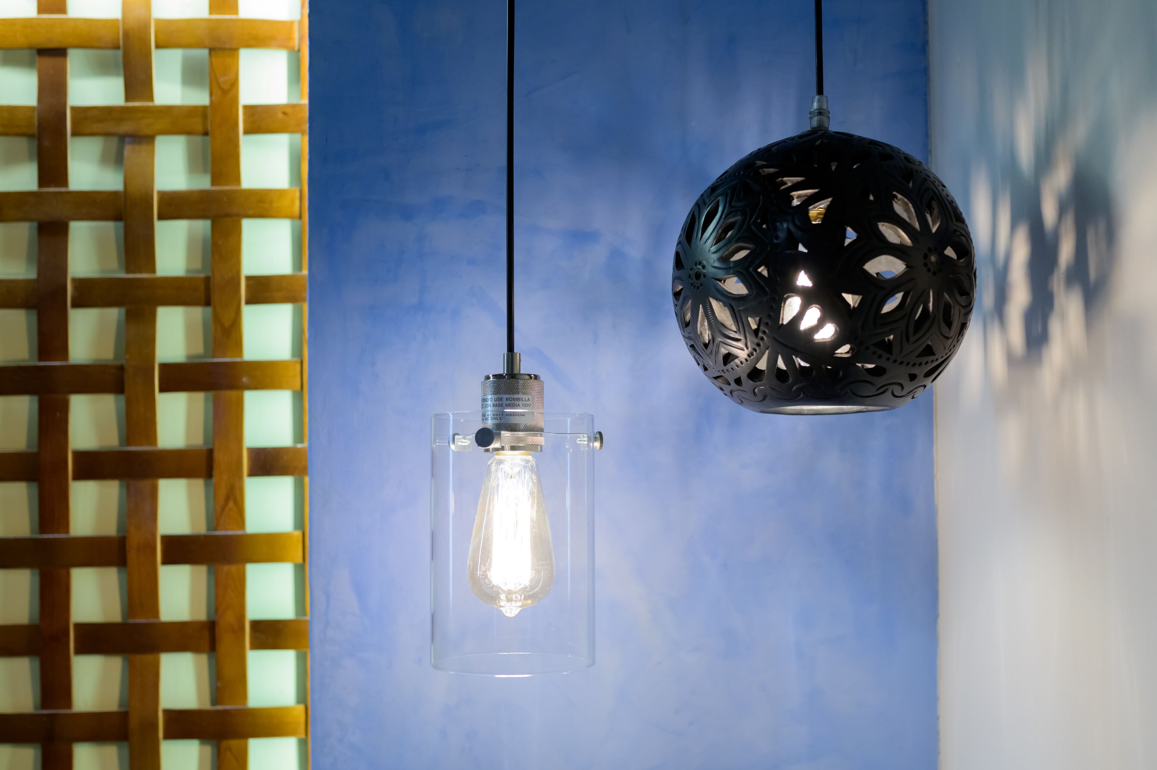 Two hanging light fixtures, one with a clear glass enclosure and one with a black perforated design, against a blue wall.