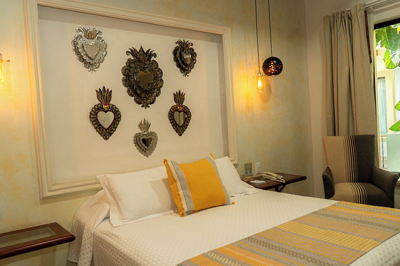 Habitación de hotel con una cama grande, una silla, una mesita de noche y una pared decorada con espejos con forma de corazón.