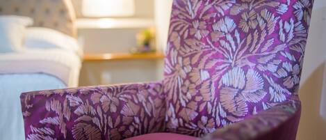 A chair with a floral pattern, a bed with white linens, and a bedside table with a lamp.