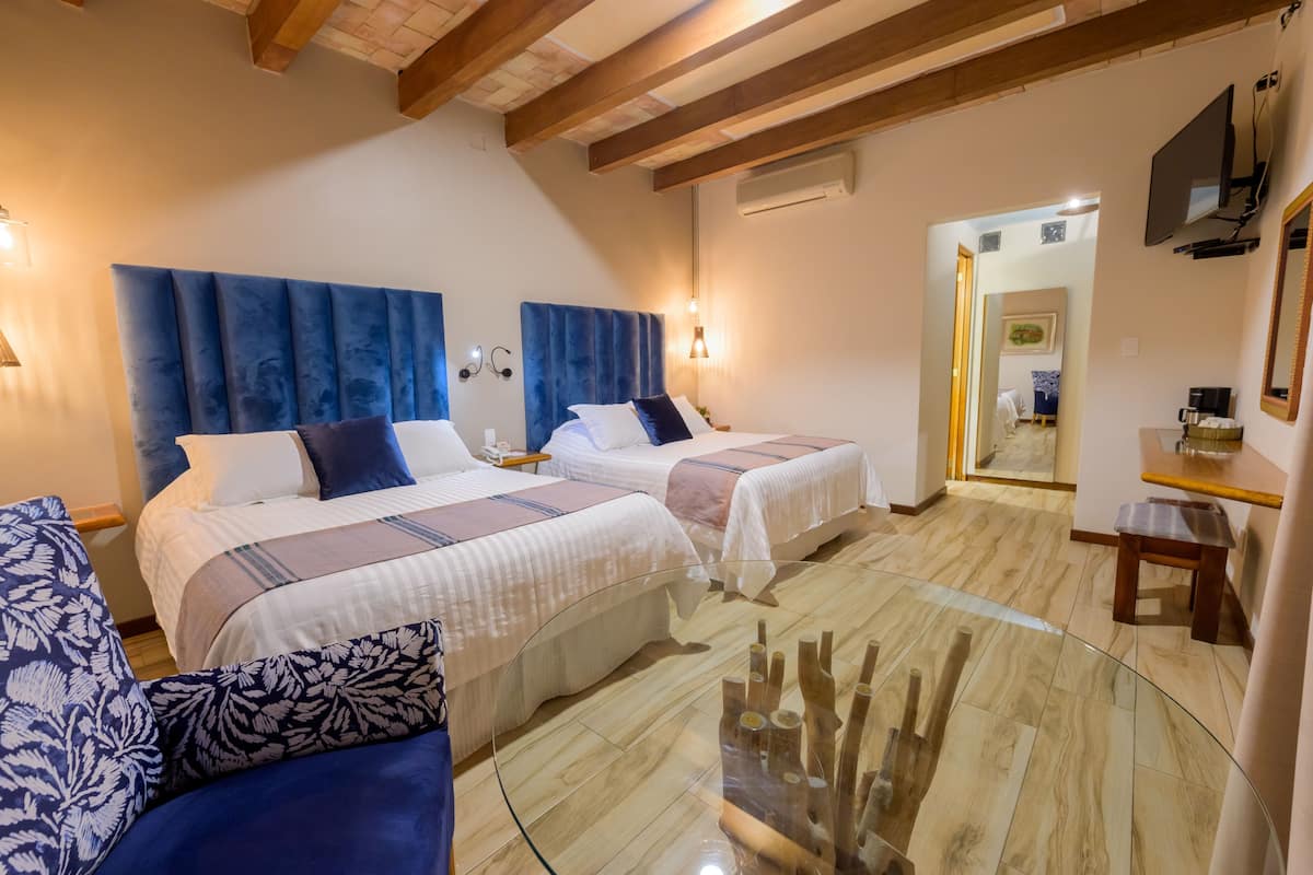 Habitación de hotel con dos camas, una mesa de centro de vidrio, vigas de madera en el techo y un televisor montado en la pared.