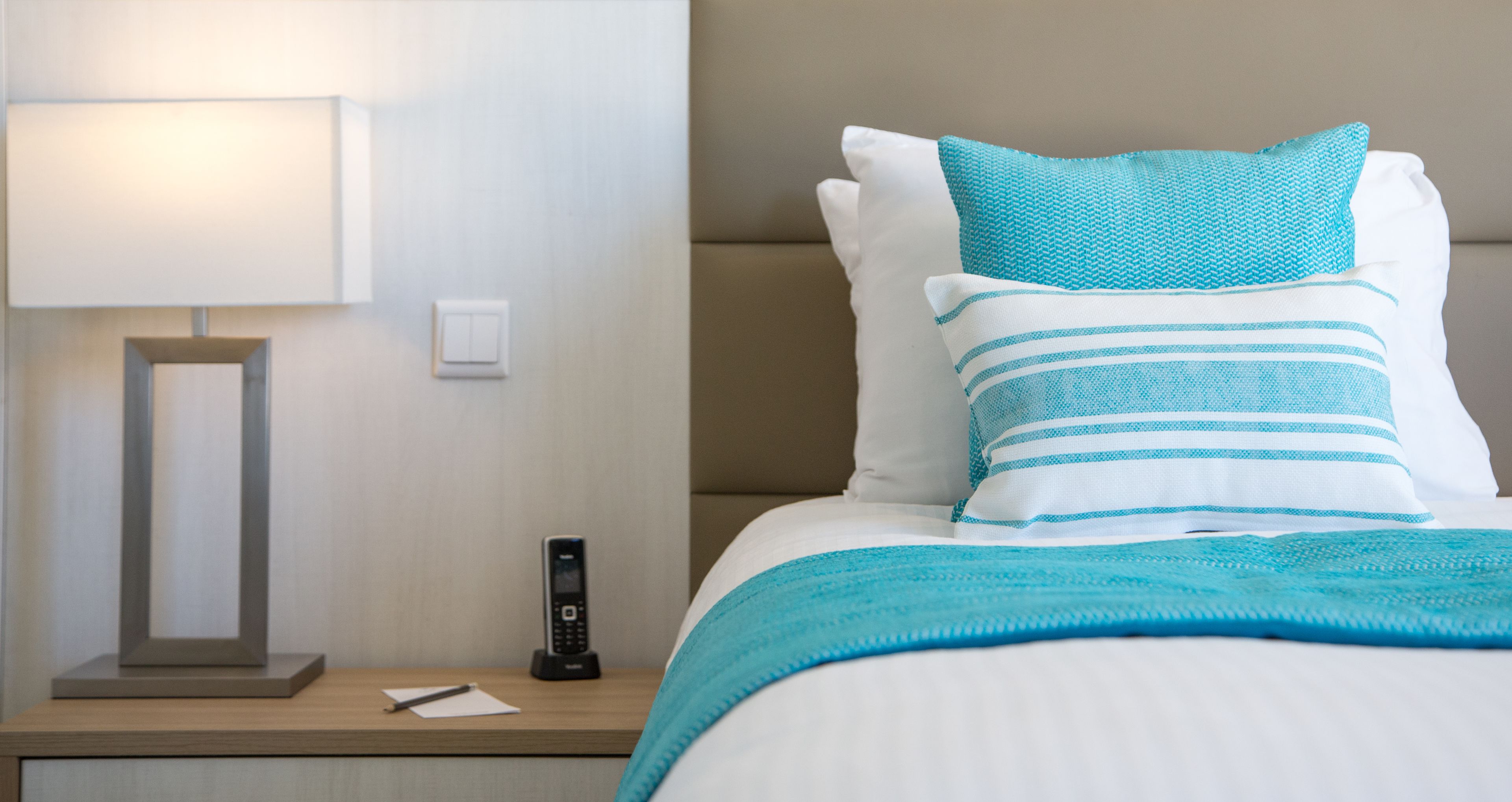 Een net opgemaakt bed met een blauw-wit gestreept kussen, een blauwgroene deken, en een nachtkastje met een lamp en een telefoon.