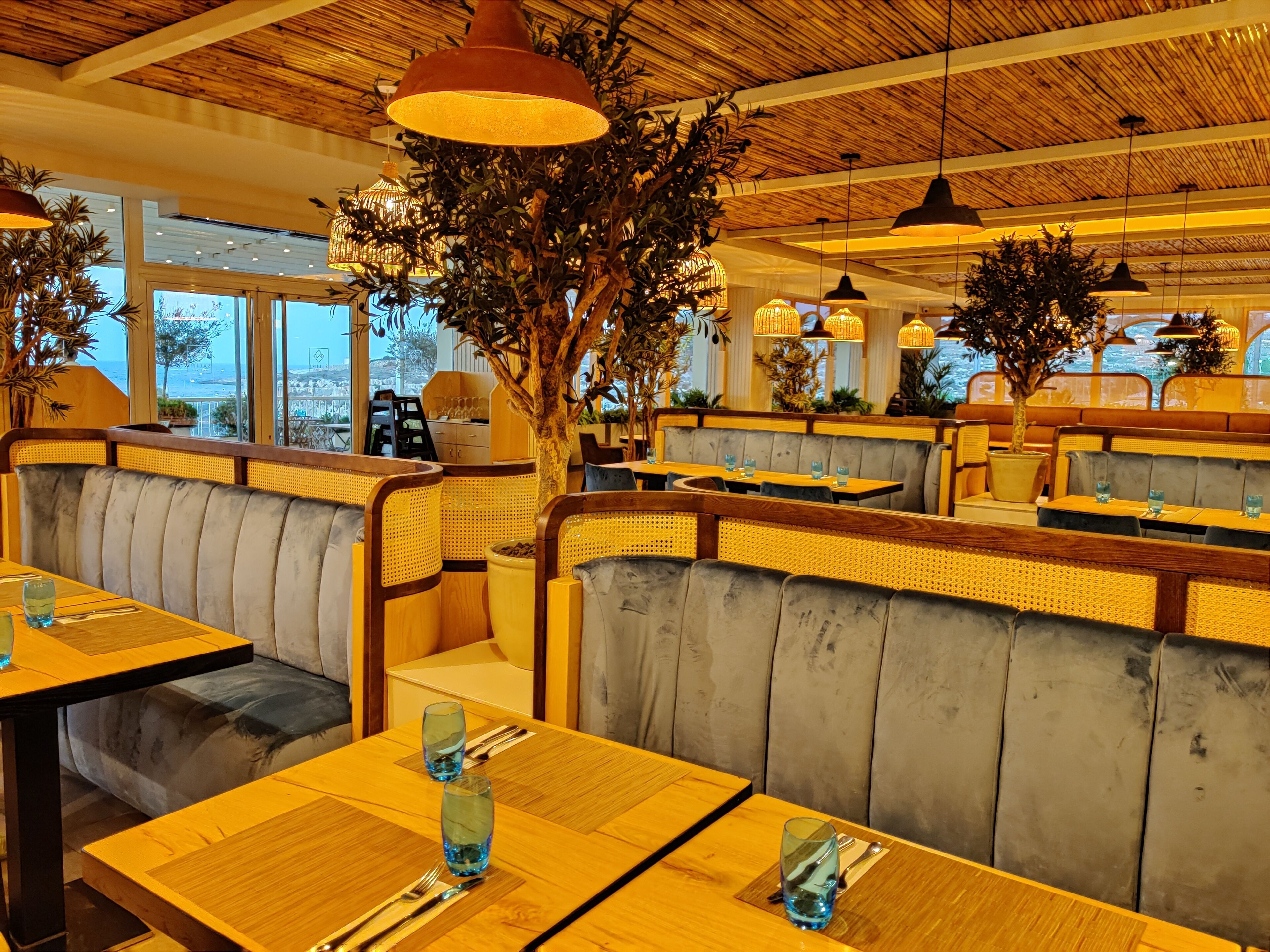 En restaurang med träbord och bekväma sittplatser, stora fönster och hängande taklampor.