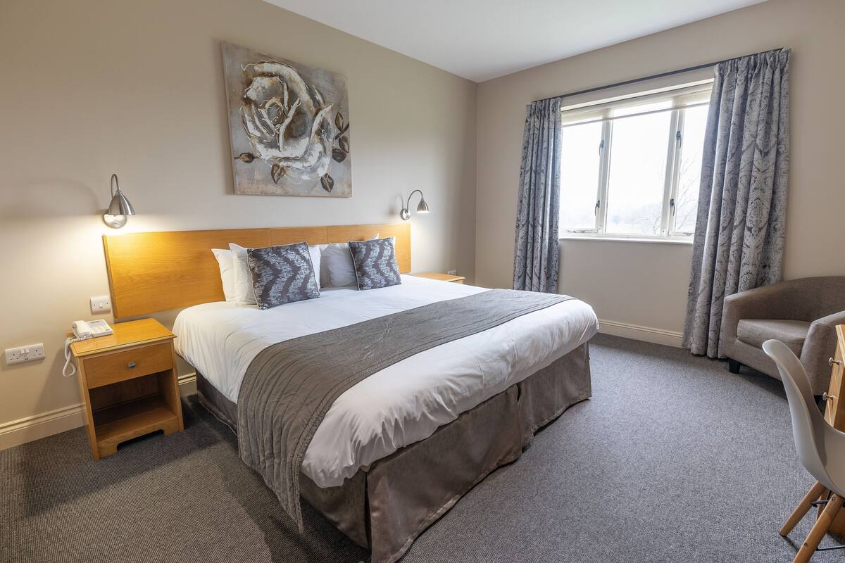 Une chambre d’hôtel avec un grand lit, des lampes de chevet, un tableau au mur, une fenêtre avec des rideaux et un fauteuil.