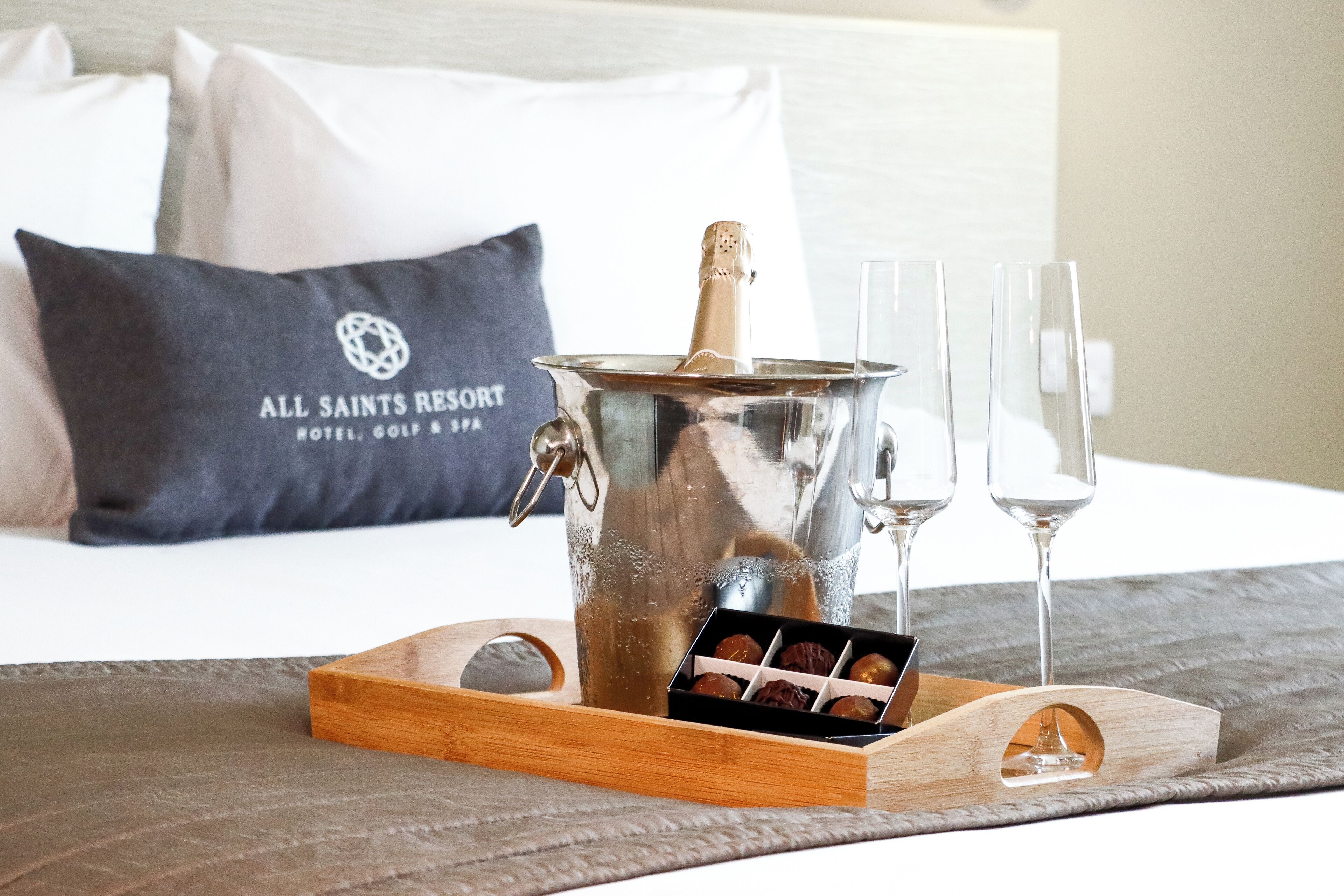 A bed with a pillow displaying the hotel name, a metal bucket with a bottle, two glasses, and a tray with chocolates.