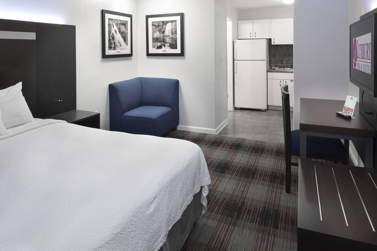 Habitación de hotel con cama, escritorio, silla, kitchenette pequeña y dos cuadros en la pared.