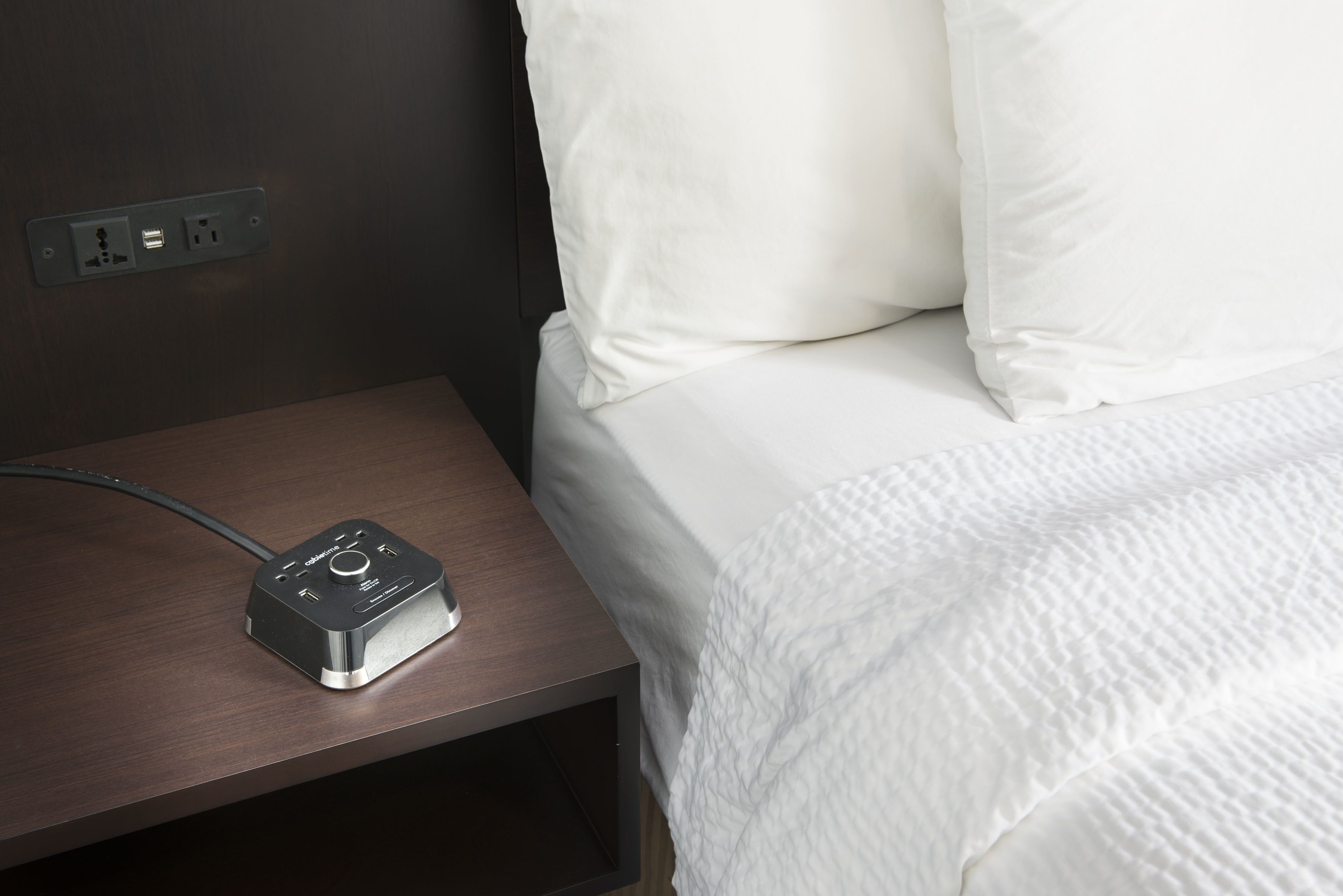 Cama bem arrumada com roupa de cama branca, mesa de cabeceira de madeira com régua de tomadas e um pequeno dispositivo eletrônico sobre a mesa.