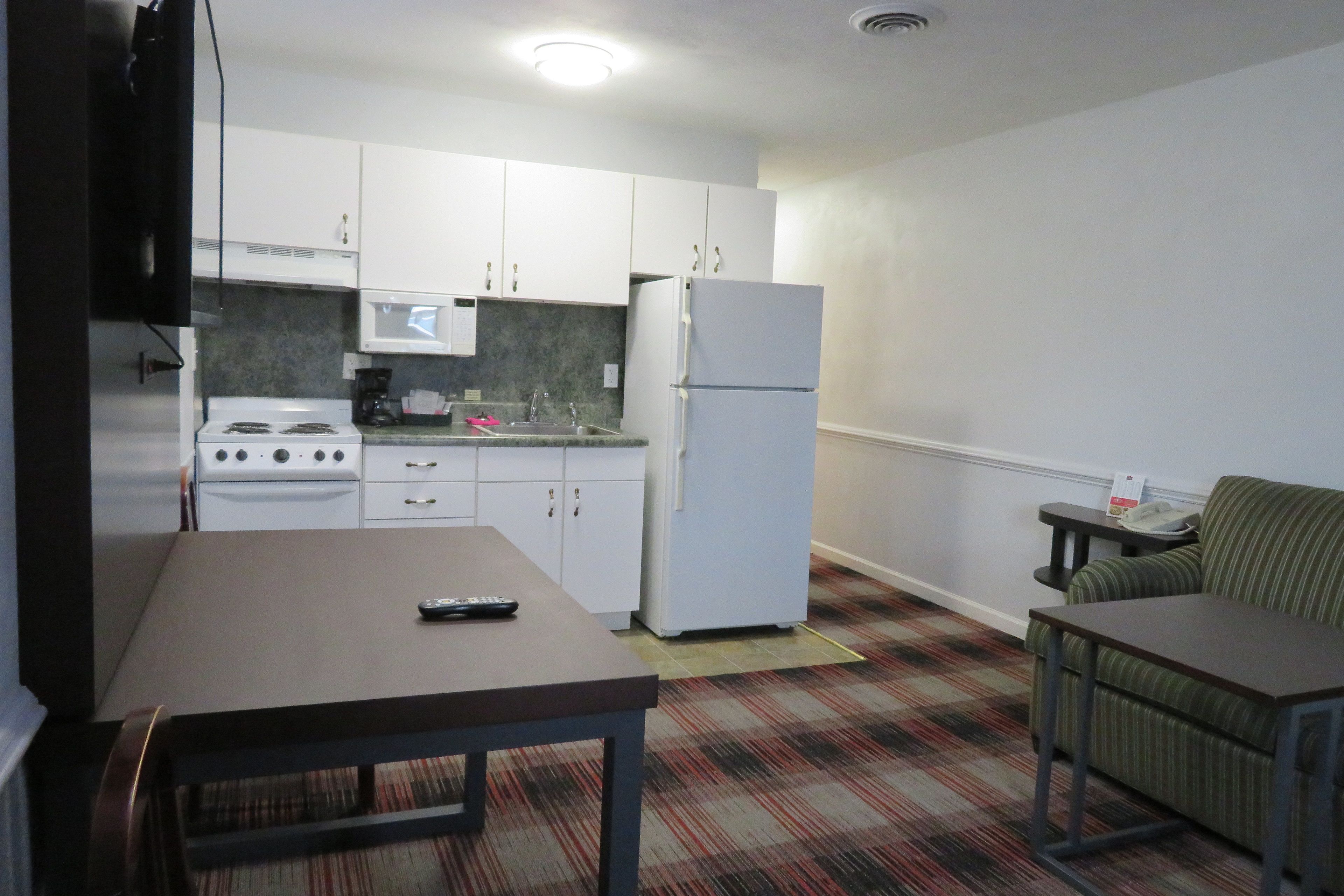 A compact kitchen with white cabinets, a stove, a microwave, and a refrigerator. There is a dining table with a remote control on it, and a striped sofa in the corner.