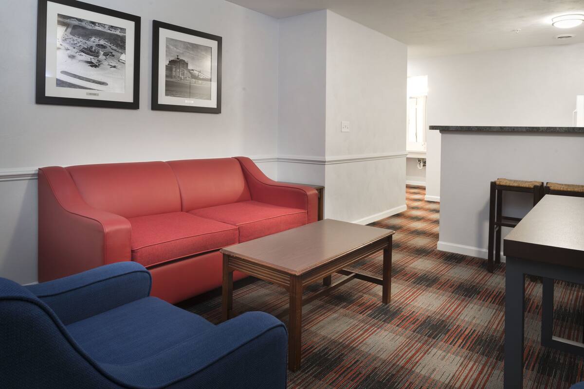 Una habitación con un sofá rojo, un sillón azul, una mesa de centro de madera y una alfombra estampada. Hay dos cuadros en la pared.