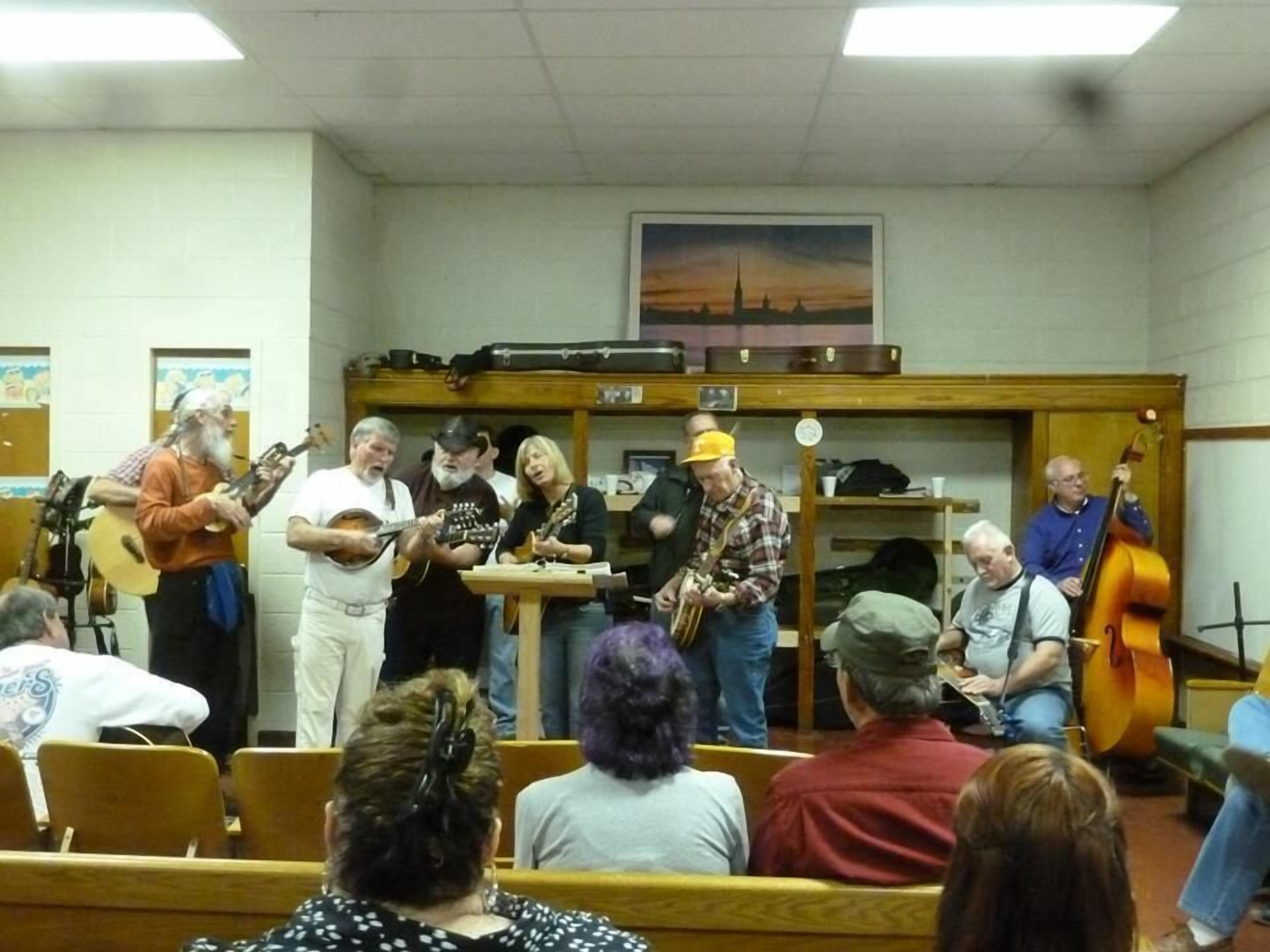 Un grupo de músicos actuando en una sala con bancos de madera y un cuadro en la pared.