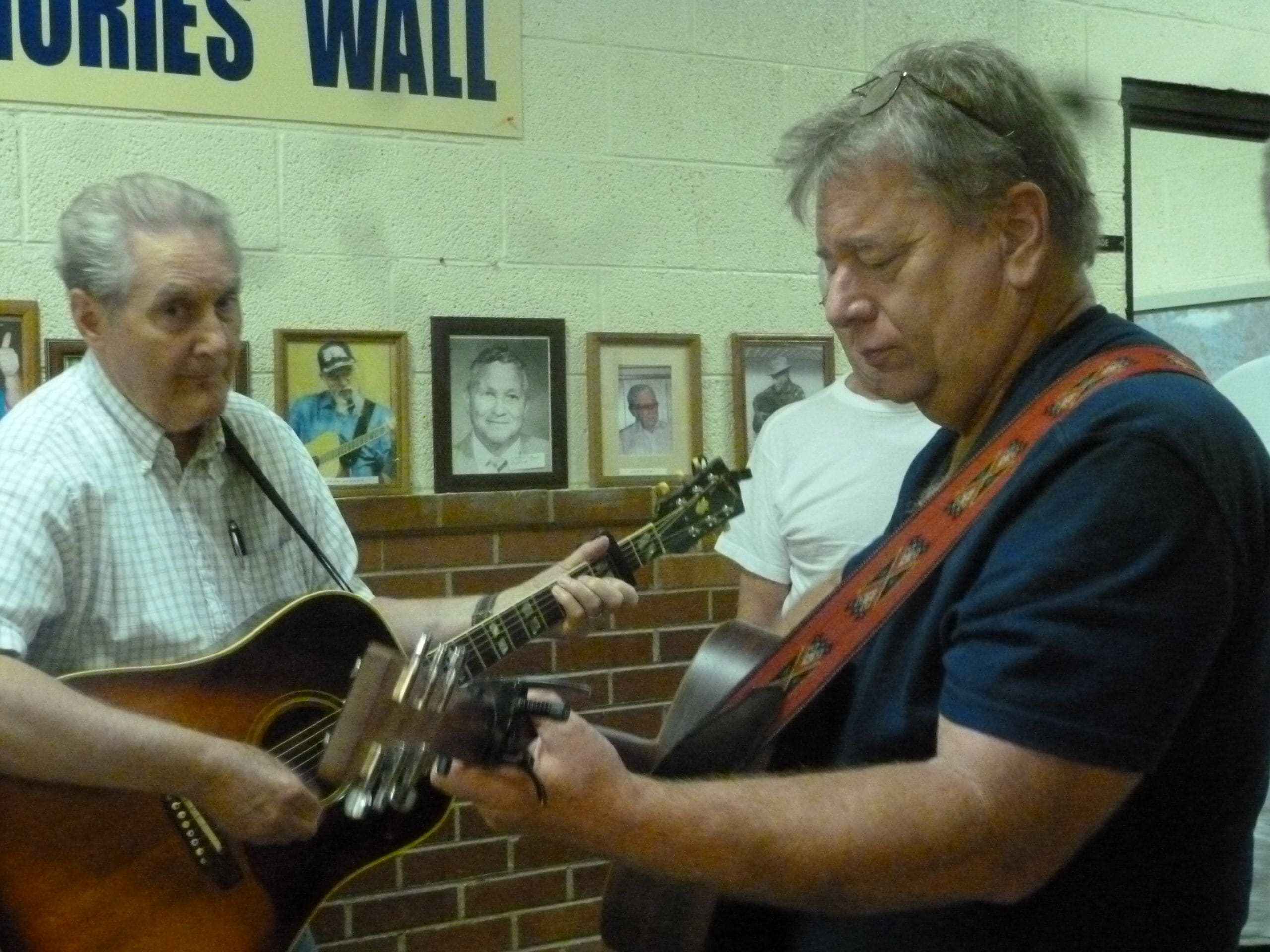 Deux hommes jouent de la guitare devant un mur orné de tableaux encadrés.