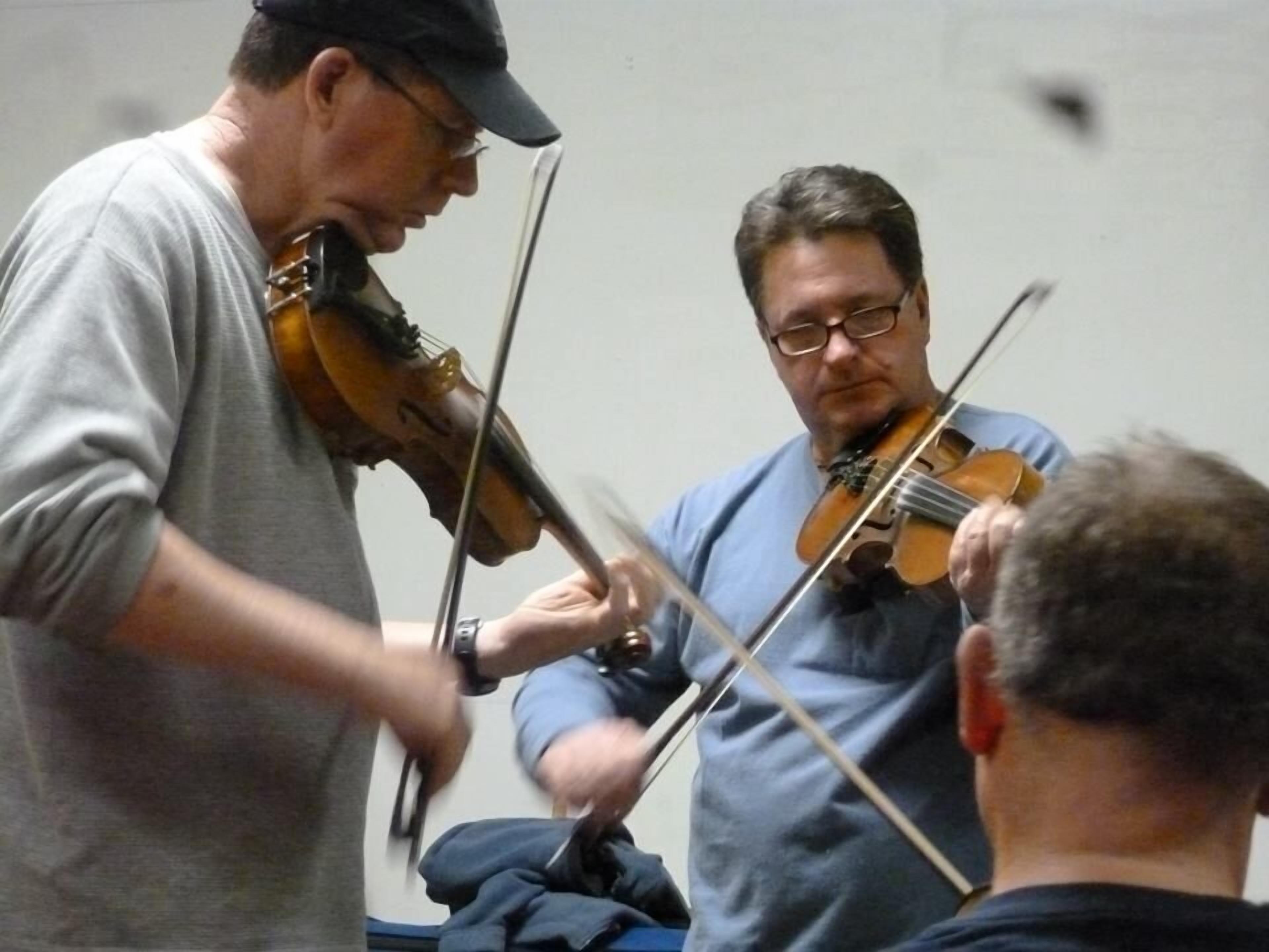 Two men playing violins in a room with a plain wall and a visible bed.