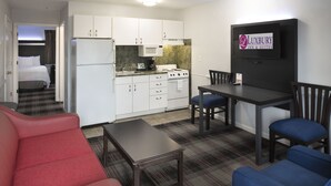 A compact hotel room with a kitchenette, a dining area with a table and chairs, a bed, and a red and blue sofa. - Luxbury Inn & Suites (Maryville)