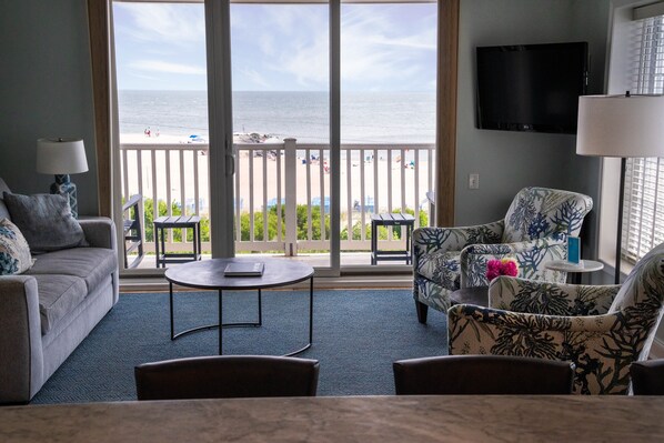 A living room with a view of the beach, a flat-screen TV, and a balcony with a railing.