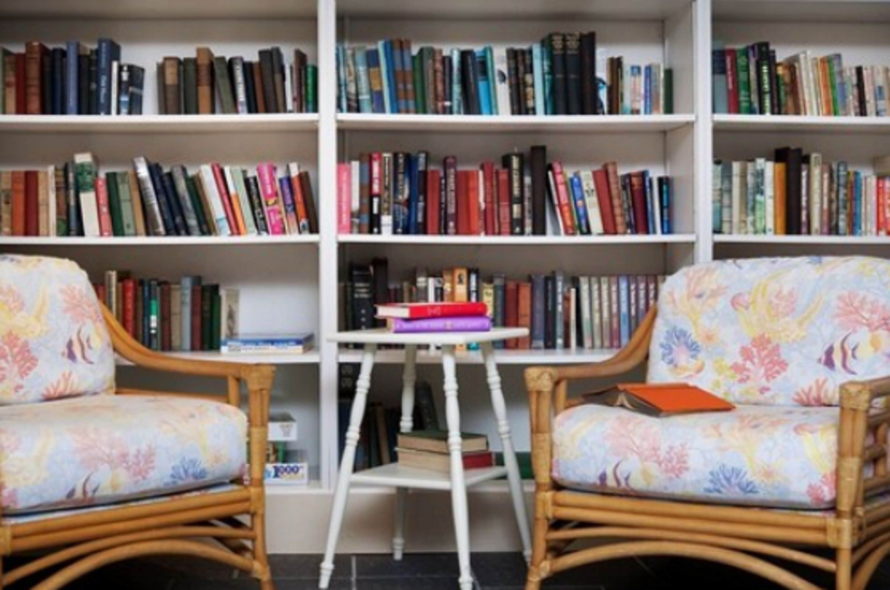A room with two wicker chairs, a small table, and a bookshelf filled with books.