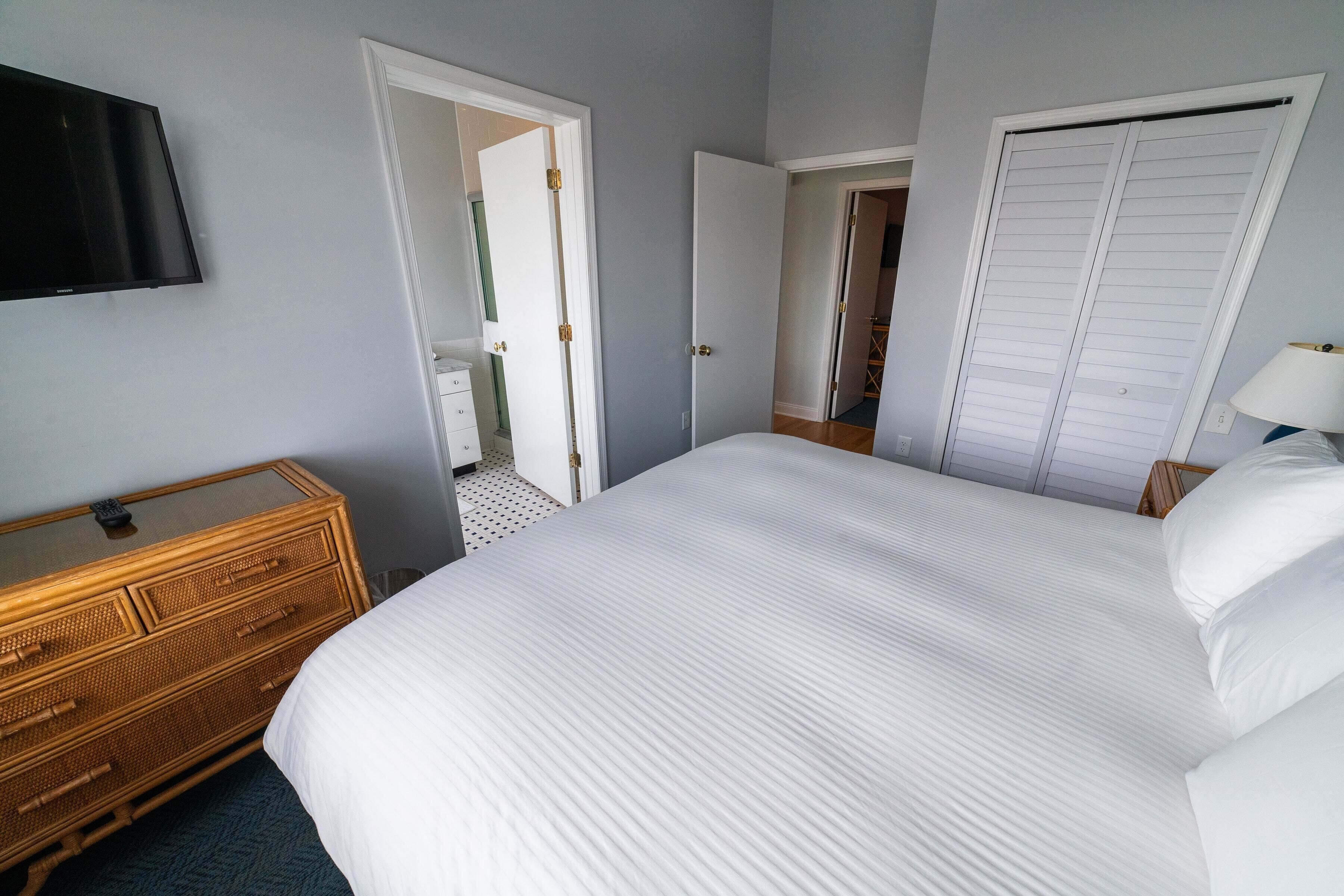 Une chambre avec un grand lit, une commode, un téléviseur fixé au mur et un placard avec des portes coulissantes.