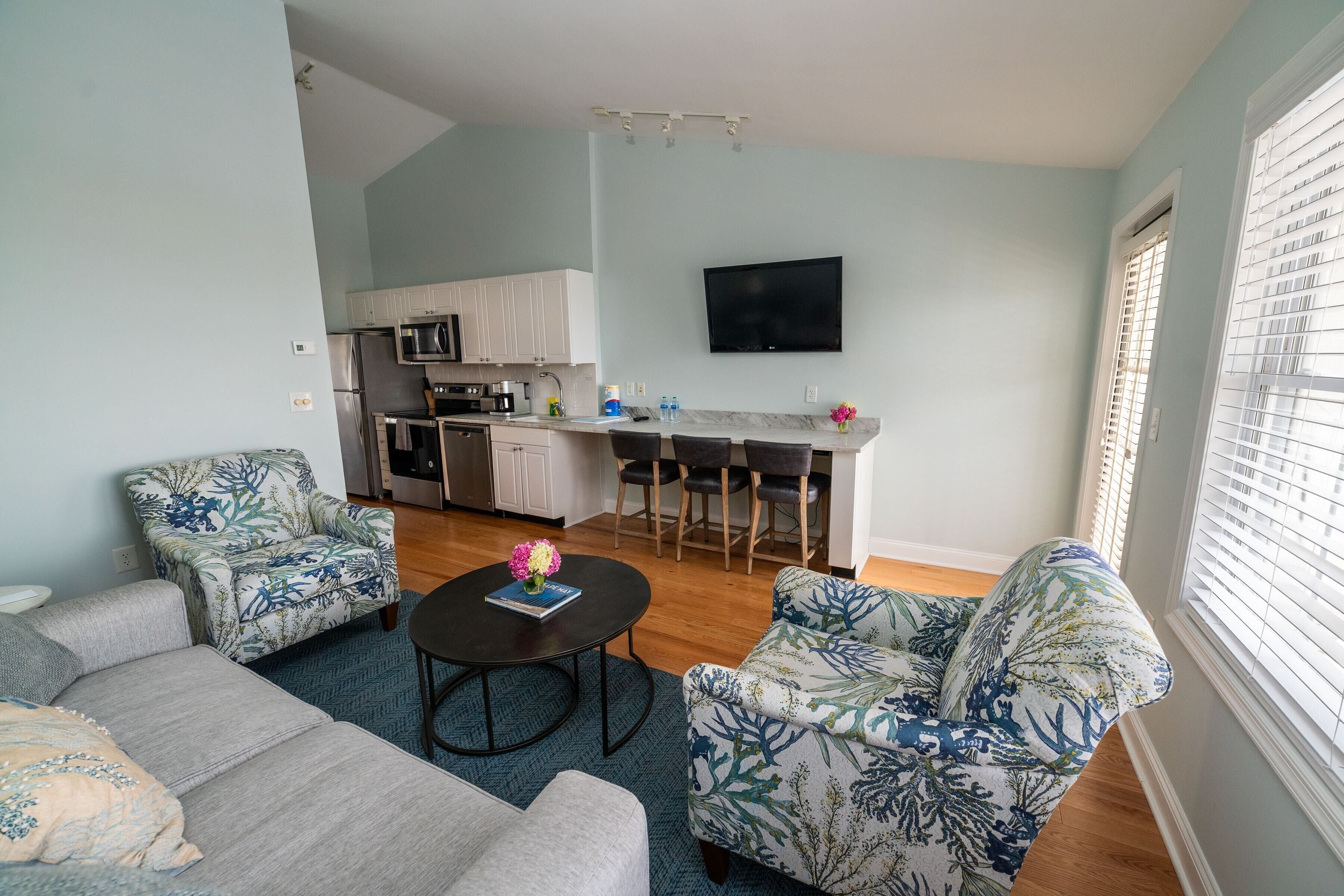 Ein modernes Wohnzimmer mit Sofa, Sessel und Couchtisch. Im Hintergrund ist eine Küche mit Essbereich zu sehen.