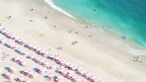 An aerial view of a beach with rows of pink and white umbrellas and beach chairs, and people enjoying the beach and the ocean.