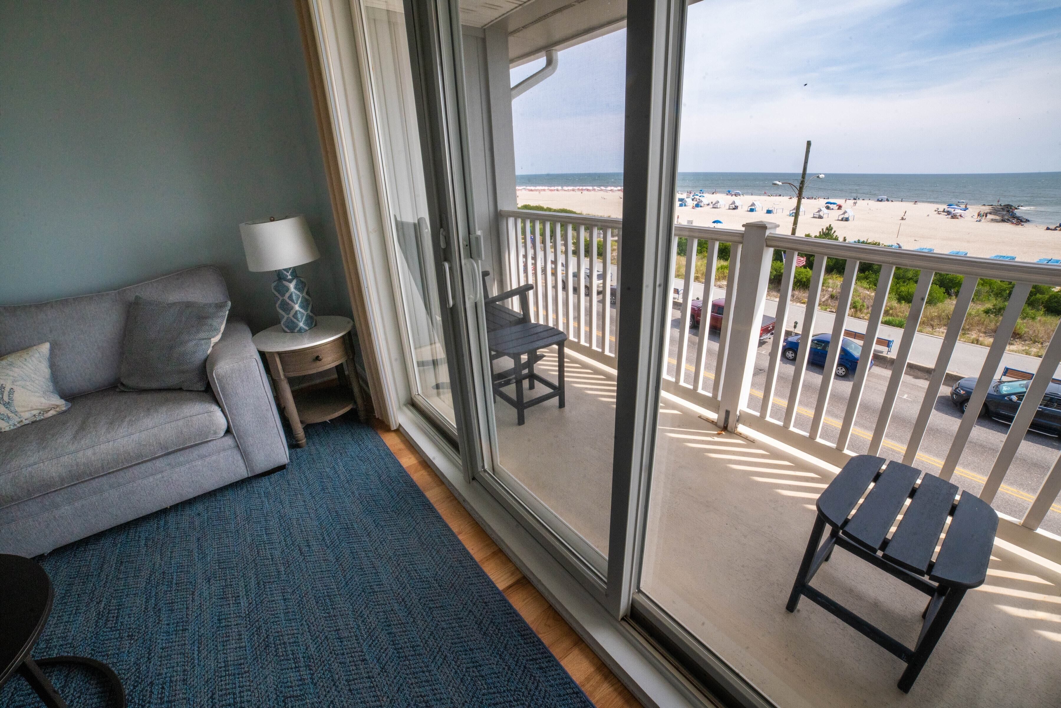 A balcony with a view of the beach, a grey sofa, a small table with a lamp, and a blue carpet.