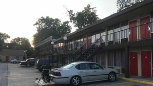 A multi-story building with a parking lot. There are several cars parked, including a white sedan and a black SUV. A bicycle is parked on the sidewalk. The building has red doors and a balcony with railings. - Budget Host Village Inn (Kirksville)