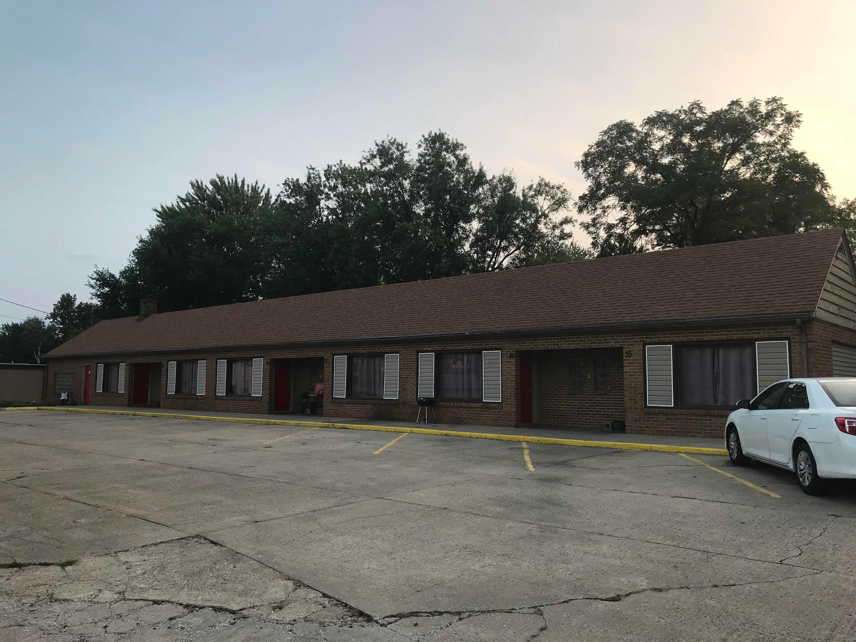 A single-story motel with a brick facade, multiple windows with shutters, and a parking lot with a white car parked in front.