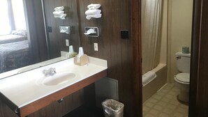 A bathroom with a wooden vanity, a sink, a mirror, and a shower area. - Budget Host Village Inn (Kirksville)