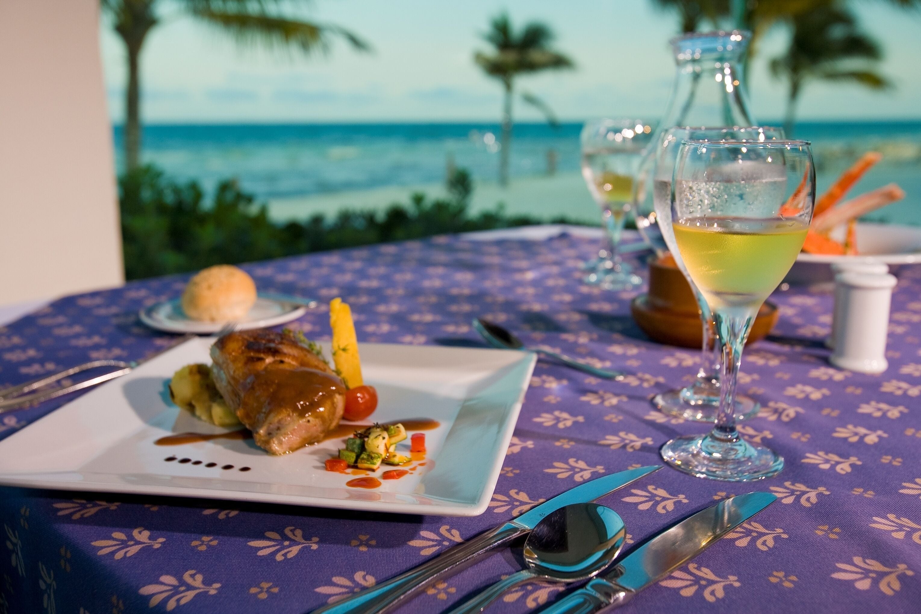 A dining table with a plate of food, a glass of wine, and a view of the ocean.