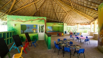 Un espacio interior amplio con techo de paja, murales coloridos y varias mesas y sillas dispuestas para comer o socializar.