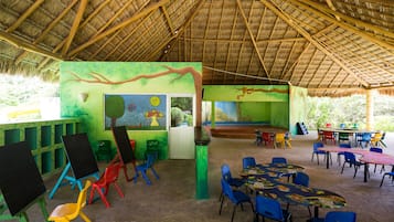 Espaço interno com telhado de palha, paredes verdes e murais coloridos. Há mesas e cadeiras dispostas para refeições ou reuniões.