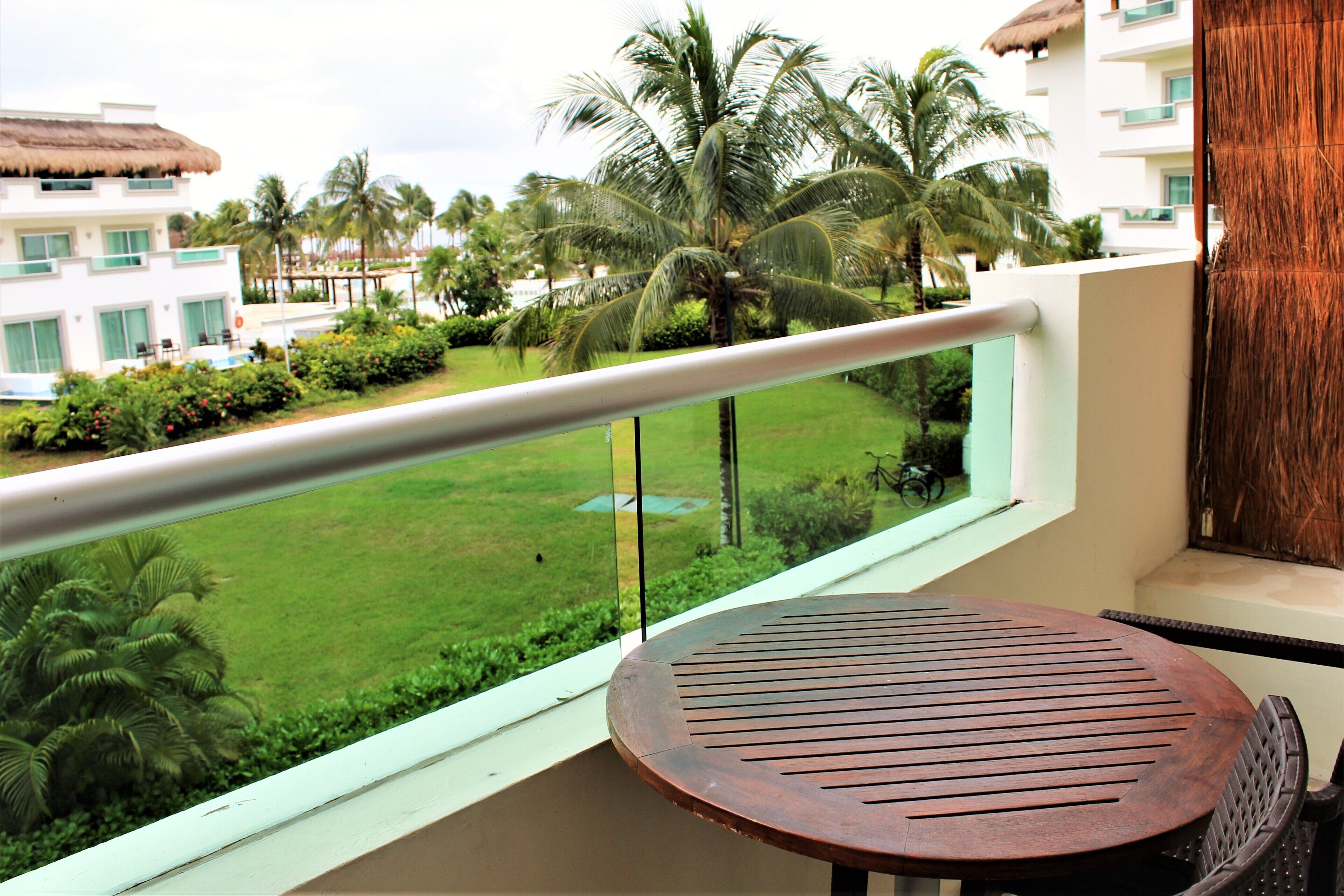 A balcony with a wooden table and a view of a well-maintained lawn, palm trees, and a building with a thatched roof.