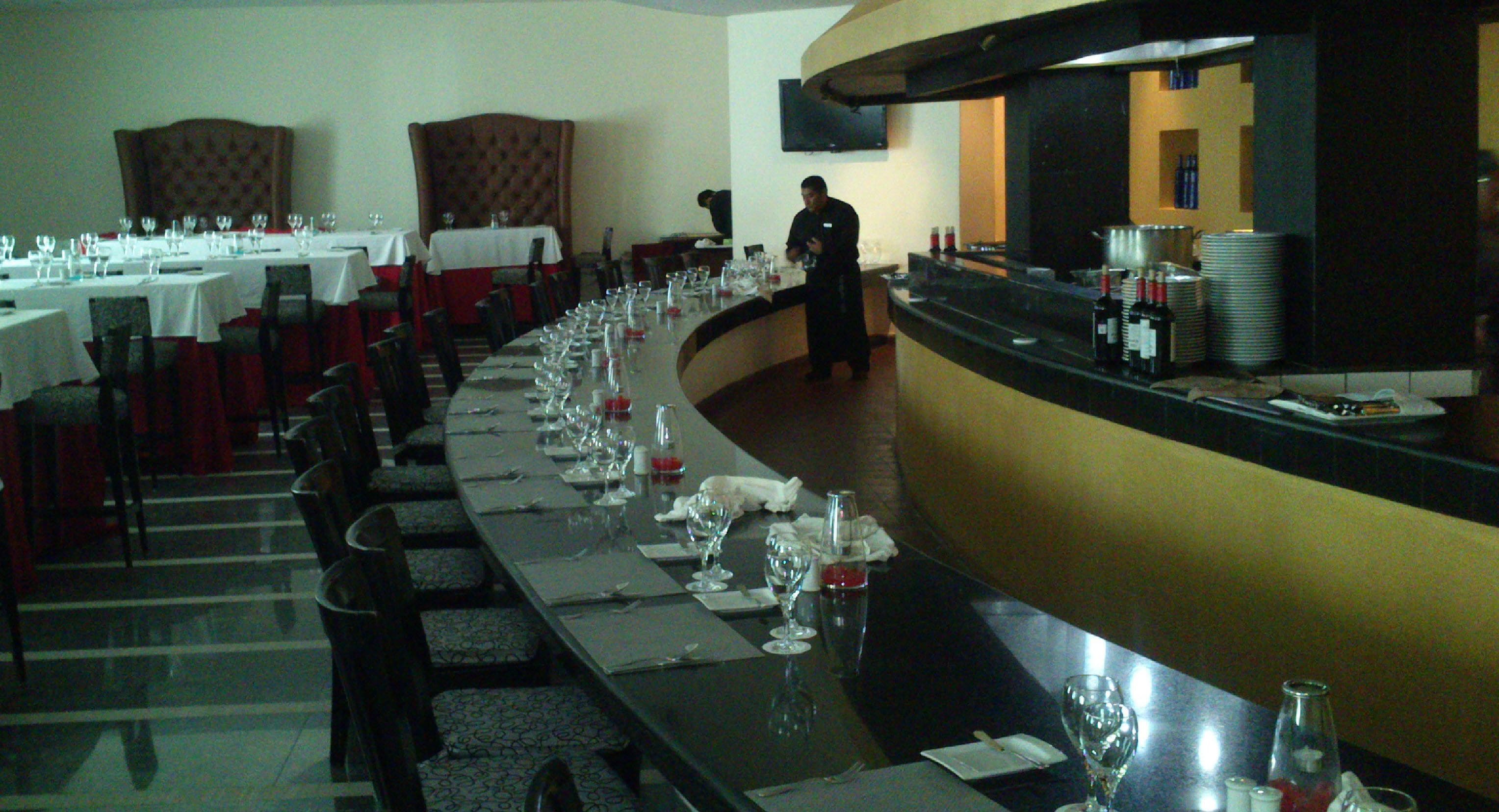 A long dining table set with multiple chairs, a bar area with bottles, and a television mounted on the wall.