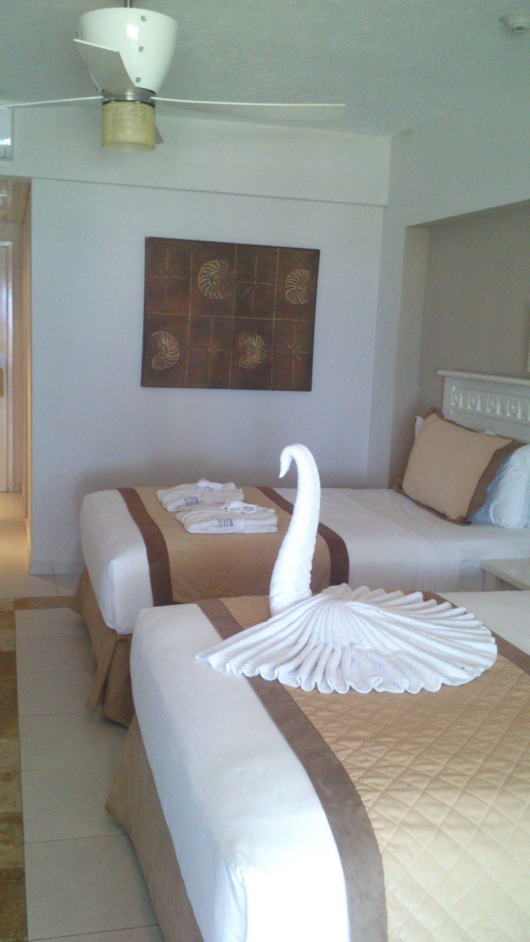 Habitación de hotel con dos camas, un ventilador de techo, una obra de arte en la pared y una cama bien hecha con toalla con forma de cisne.