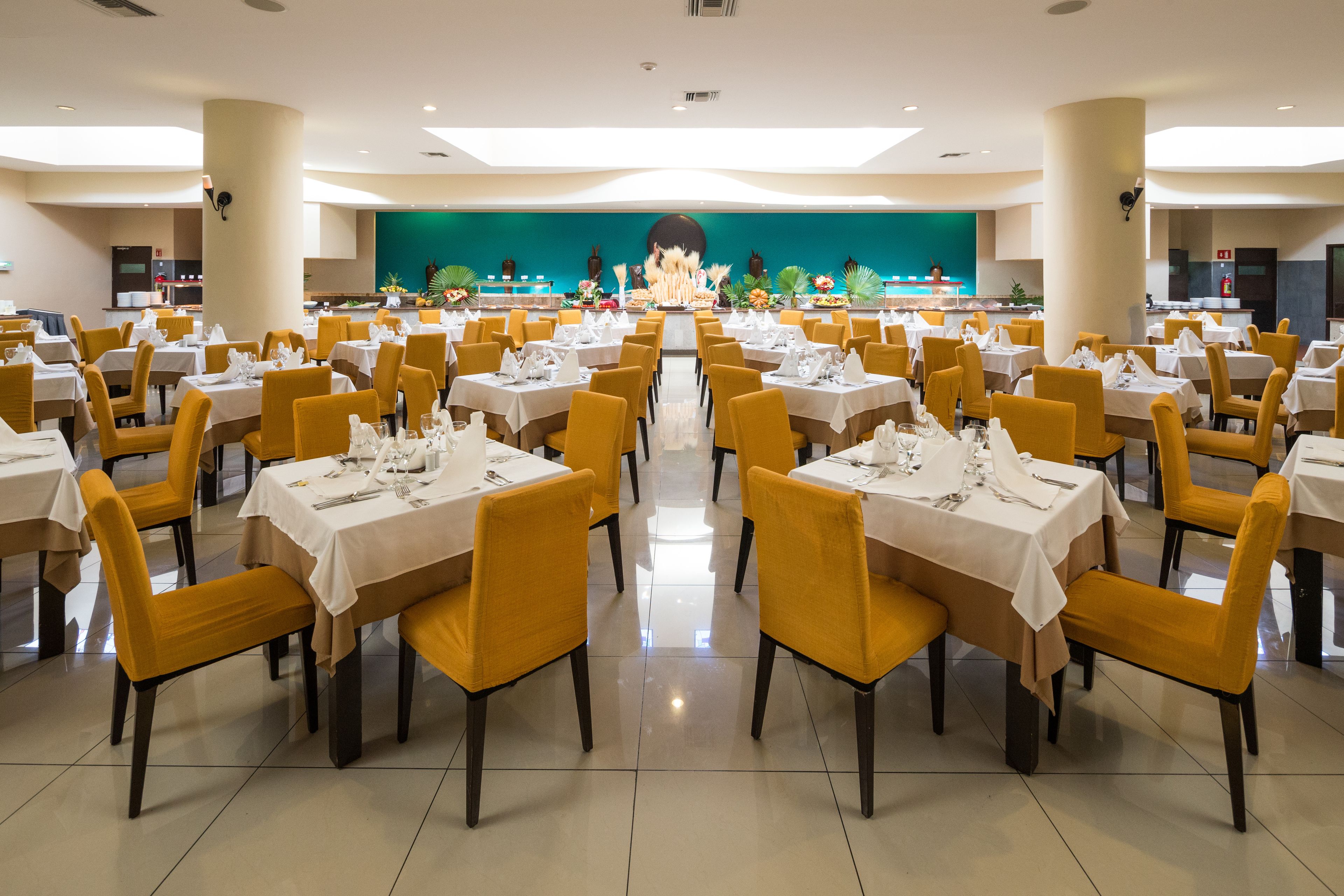 A large dining hall with multiple tables and chairs, a stage with a large screen, and a green backdrop.