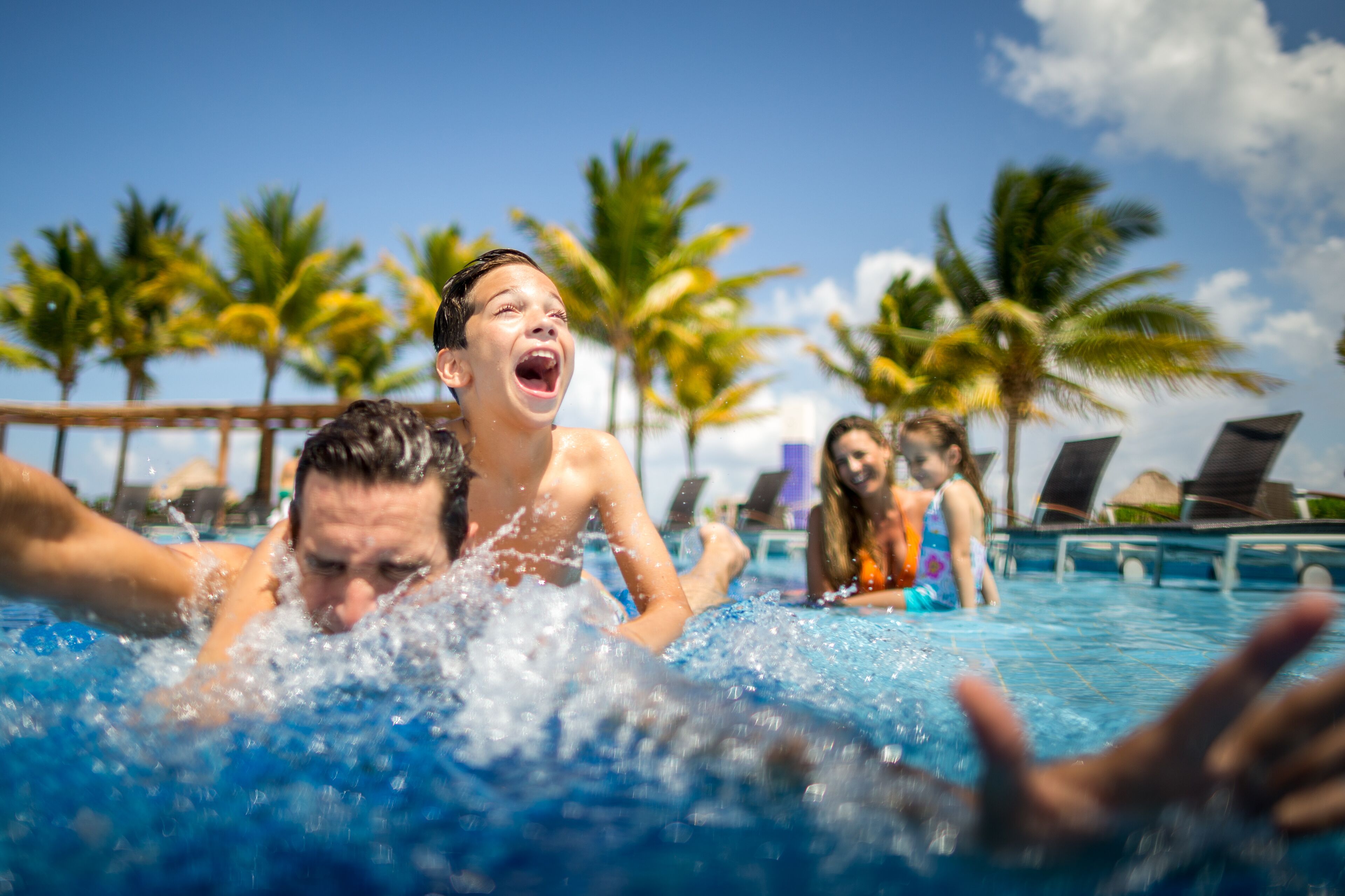Ein Schwimmbecken mit Personen im Wasser, Palmen und Liegestühlen im Hintergrund.